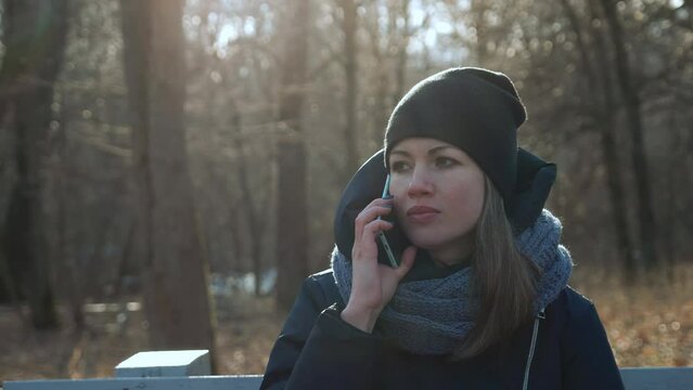 Young white girl sitting on a park bench wearing a coat, talking on the phone and smiling. Beautiful caucasian woman sits in the city park in early spring or late autumn. full HD 50p video footage