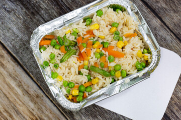 Portion of rice with mixed vegetables in a foil takeaway container and lid.  On a wooden background