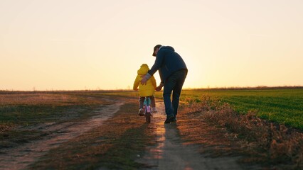Dad teaches child to ride bike in park at sunset, happy family. Father teaches kid, girl, daughter to ride childrens bike on road in spring. Happy childhood, childhood dreams. Dad kid walking together