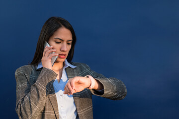 Latin businesswoman looking at her smartwatch and talking on cellphone on blue background.