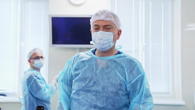 Adult male doctor in mask speaks to the camera. Surgeon at backdrop tuning the monitor before the operation in blur.