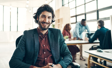 Greet every workday with a positive attitude. Portrait of a young businessman sitting in a modern office with his colleagues working in the background.