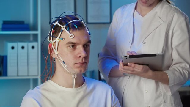 EEG topographer for examining brain, doctor making electrogram of scalp activity. Researching of brain work man wearing electroencephalography headset, nurse writing notes in gadget tablet.