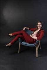 Cute plus-size model is sitting on a chair on a black background in an unbuttoned red jacket, a pink blouse, black trousers and high-heeled shoes. Business portrait. Business woman in red suit