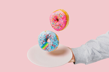 Two multi-colored donuts hover over a plate held by a hand on a pink background. Unhealthy food...
