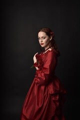 Fototapeta premium portrait of pretty female model with red hair wearing glamorous historical victorian red ballgown. Posing with a moody dark background.