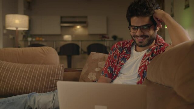 Track in shot of a young happy Indian Asian attractive male or man wearing specs sitting on a couch and smiling while watching a Comedy Movie or video on Ott platform using a laptop at home night