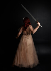  Full length portrait of pretty female model with red hair wearing glamorous fantasy tulle gown and crown.  Posing with a moody dark background.