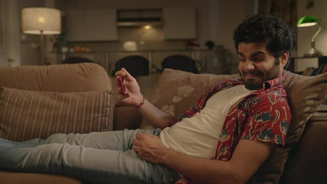 A young modern Asian Indian man is trying to watch a horror or violent movie or film on an OTT platform using mobile phone is scared afraid to look at the display or screen in an interior night home