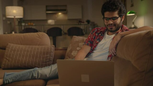 Track out shot of a smiling young happy Indian Asian attractive male or man wearing specs sitting on a couch and smiling while watching a Movie or video on Ott platform using a laptop at home night