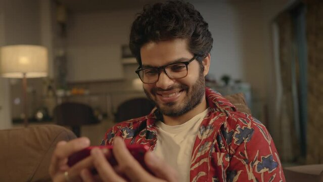 close up shot of a young happy Indian Asian attractive male guy wearing specs sitting on a couch and smiling while watching a Comedy Movie or video on Ott platform using a mobile phone at home night