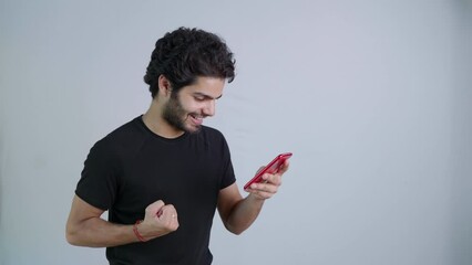 A young Indian Asian excitedly happy male or man holding a mobile phone and celebrating a lottery win, betting or winning game through a online fantasy sports platform isolated on white background.