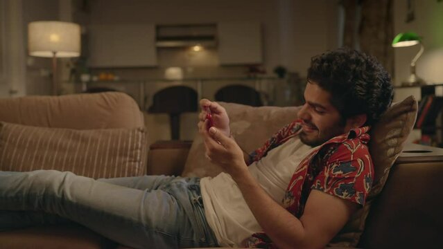 A young modern happy Asian Indian man or male is resting on a couch laughing and watching an online funny or comedy movie or video on an OTT platform in an interior night house setup.