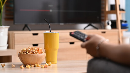 Cropped shot bowl of popcorn and glass on wooden table with young man holding remote control while watching TV in living room.