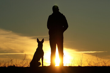 Silhouettes of a man and a dog on a sunset background, the owner walks with his pet in nature,...