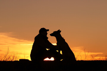 Silhouettes of a man and a dog on a sunset background, the owner walks with his pet in nature,...