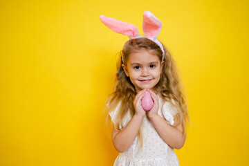 beautiful smiling blonde girl with bunny ears holding an easter egg in her hands on a yellow background, looking at the camera, kid celebrate easter.