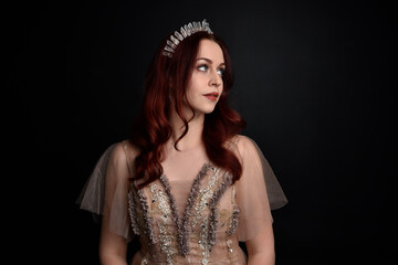 portrait of pretty female model with red hair wearing glamorous fantasy tulle gown and crown.  Posing with a moody dark background.