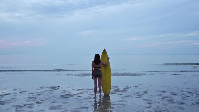 Young asian woman in swimsuit with yellow surfboard standing on the beach