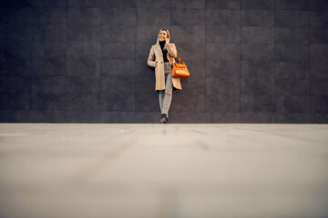 A trendy woman in a coat leaning on the wall outside and talking on the phone.