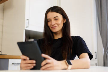 Cheerful white woman browsing internet on laptop computer in home kitchen. Happy brunette female with long dark hair using modern device for online communication and entertainment during lockdown