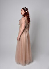 Fototapeta premium Full length portrait of pretty female model with red hair wearing glamorous fantasy tulle gown and crown. Posing with gestural arms on a studio background