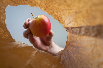 A hand places a red apple in a brown paper bag. A mature man's hand holds the ripe fruit over the...