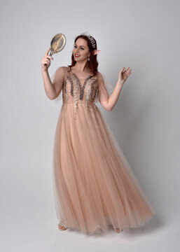 portrait of pretty female model with red hair wearing glamorous fantasy tulle gown and crown.  Posing with gestural arms  holding a hand mirror on a studio background