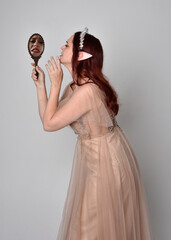 portrait of pretty female model with red hair wearing glamorous fantasy tulle gown and crown.  Posing with gestural arms  holding a hand mirror on a studio background © faestock