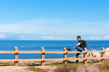 Man with backpack sitting on the fence at the end of earth watching on bright blue lake. Hiking or travel concept. Exploring the great outdoors. Beauty of nature