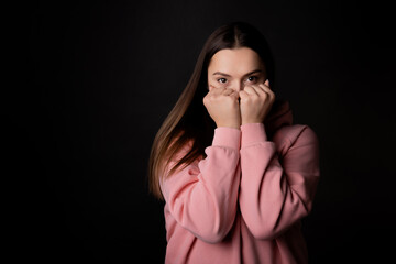 A cute young brunette in a pink hoodie hides her face with her hands, portrait on a black background.