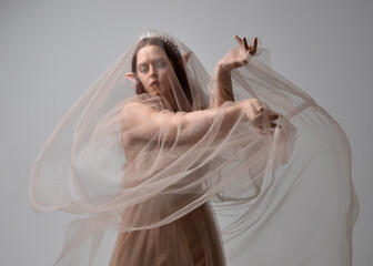  Full length portrait of pretty female model with red hair wearing glamorous fantasy tulle gown, crown and shroud veil.  Posing with gestural arms on a studio background
