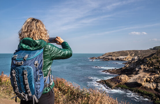 Walking along the seashore. A woman walking with a backpack taking a photo of the cliffs with her cell phone.