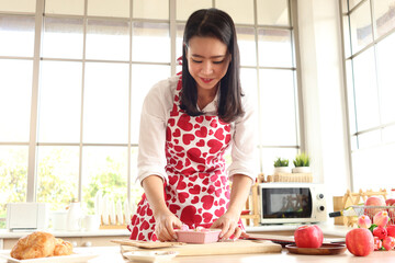 Happy smiling beautiful young Asian woman in cute red heart apron holding valentine homemade cookies, standing behind kitchen counter, preparing romantic love memorable anniversary celebration.