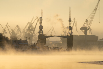 Cranes of Baltic shipyard in St. Petersburg in frosty winter day, steam over the Neva river, smooth surface of the river at sunset.