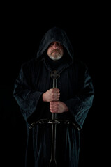 Portrait of courageous monk-warrior in black cloak with sword in hands on black background....