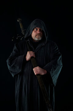 Portrait of courageous monk-warrior in black cloak with sword in hands on black background. Historical medieval templier fantasy