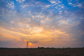The sky is orange and blue in the evening with white clouds and the sunset. the silhouette of the high voltage power pole