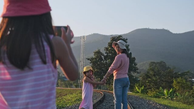 A sister takes a picture of mother and small sister with smartphone to record memories while walking on railroad tracks in the countryside against the mountains in the evening.
