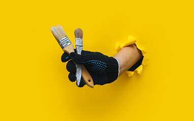 The right painter's hand in a black cloth knitted glove holds a two bristle brush. Torn hole in yellow paper. The concept of a worker, a labor migrant, a master of his craft. Copy space.