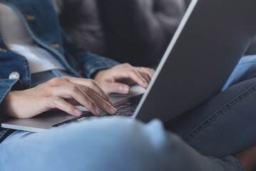 Fototapeta premium Young woman sitting on sofa, working and typing on laptop computer at home, working at home, telecommuting concept