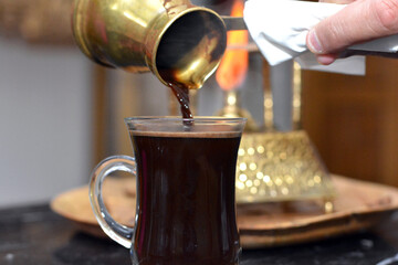 Pouring Turkish coffee in a glass mug from a copper pot made on the flame of a handmade engraved...