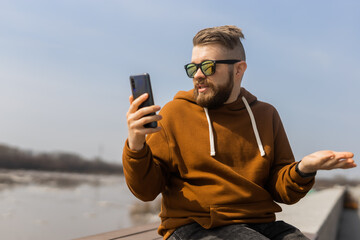 Millennial bearded man making video call on mobile phone outdoors in city. Videocall smartphone and technologies concept