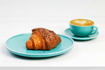 Croissant with chocolate and cup of coffee cappuccino on white wooden table.