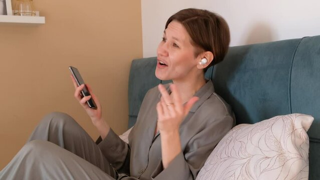 Happy woman in earphones singing and listening to music while sitting on bed and holding mobile phone at home.