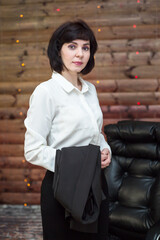 brunette woman in a white blouse with a black jacket on her arm against a wooden wall with a garland.