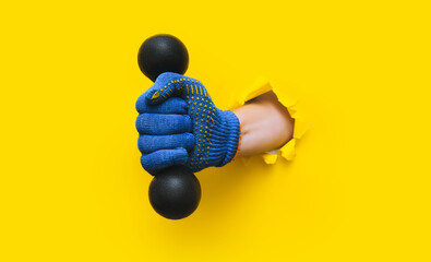 A worker's hand in a blue knitted glove holds a black dumbbell. Torn hole in yellow paper. Healthy lifestyle concept avd copy space.
