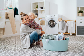 Mother sits on laundry room floor with little daughter, a cute little girl with blonde hair tied up in a bun grabs the woman by neck hugs her tightly they spend time doing housework sorting clothes. - obrazy, fototapety, plakaty