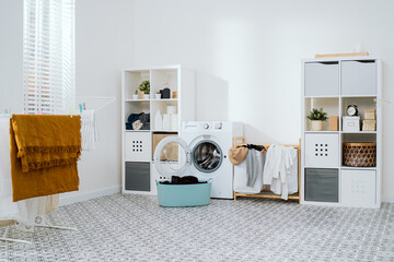View of home laundry room, dresser with softeners, powder, towels, open washing machine with empty drum, next to wicker basket with colorful laundry items, dryer - obrazy, fototapety, plakaty