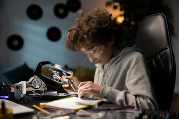 Child spends free time at desk learning to solder, cares about safety, boy has safety goggles, repairs cables, electronics, smoke rises
